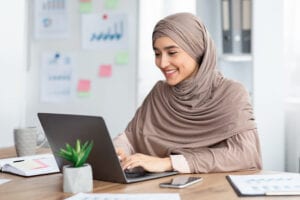 Office Job. Happy islamic woman in hijab working on laptop at desk, typing on keyboard and looking at computer screen.